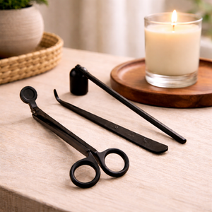Matte black wick trimmer, snuffer and dipper beside a lit candle