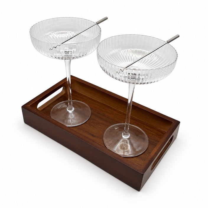 Two ribbed coupe glasses and metal cocktail picks on a rectangular wooden tray against a white background