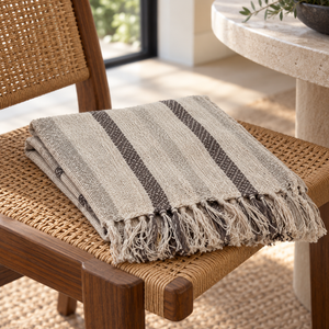 Beige striped cotton throw with fringed ends folded on a woven chair