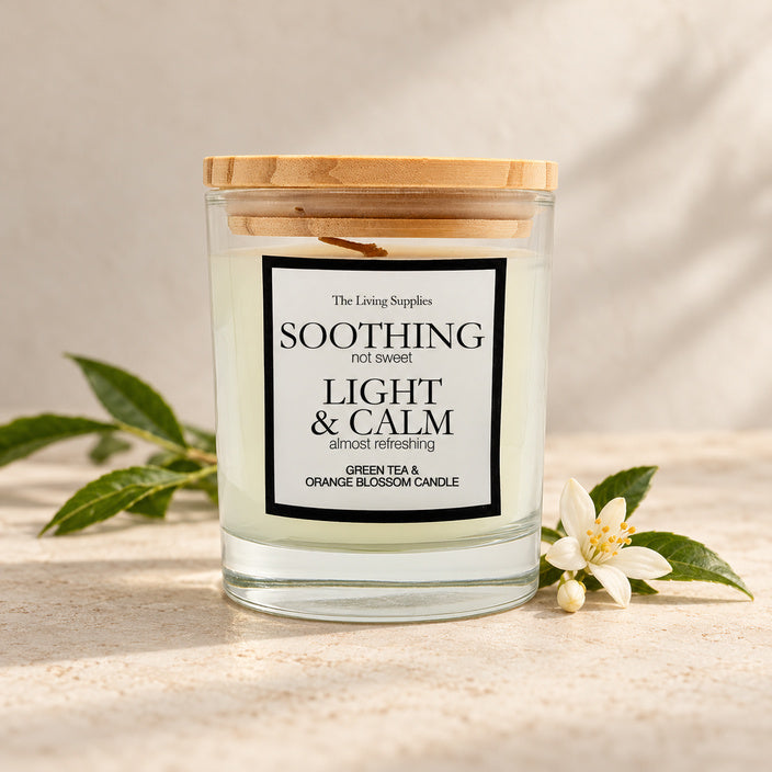 The Living Supplies Green Tea & Orange Blossom candle in a clear glass jar with a wooden lid, beside green leaves and white orange blossom.