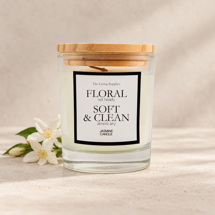 The Living Supplies Jasmine candle in a clear glass jar with a wooden lid, beside white jasmine flowers.