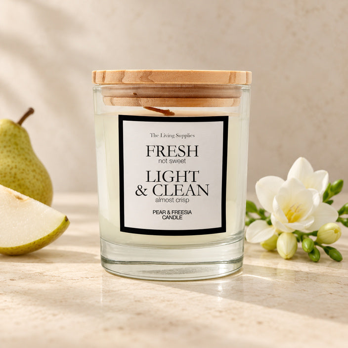 The Living Supplies Pear & Freesia candle in a clear glass jar with a wooden lid, beside pear slices and white freesia flowers.