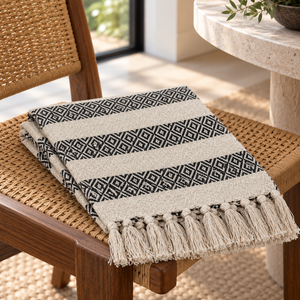 Beige Rosie cotton throw with black geometric bands and fringe folded on a woven chair