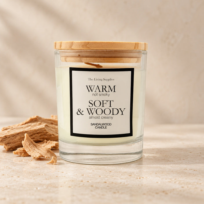 The Living Supplies Sandalwood candle in a clear glass jar with a wooden lid, beside sandalwood pieces.