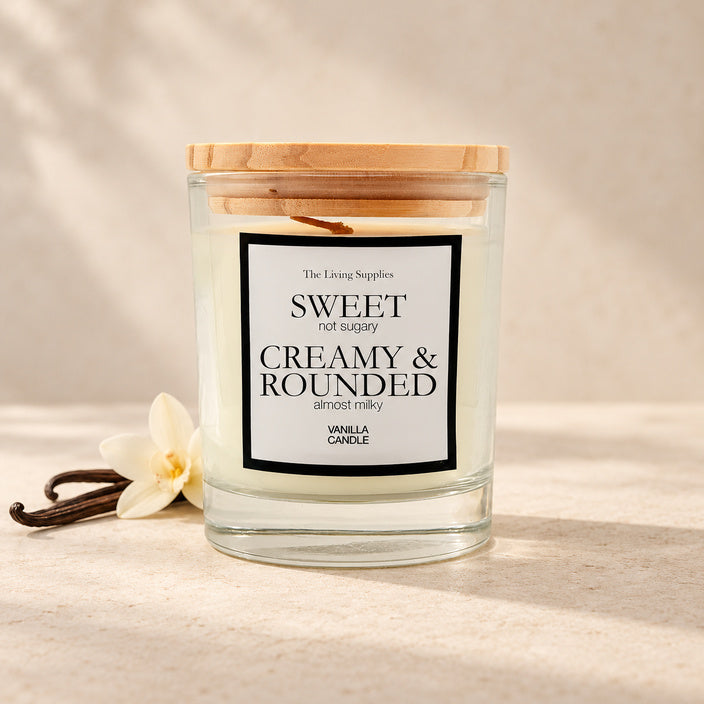 The Living Supplies Vanilla candle in a clear glass jar with a wooden lid, beside vanilla pods and a white flower.